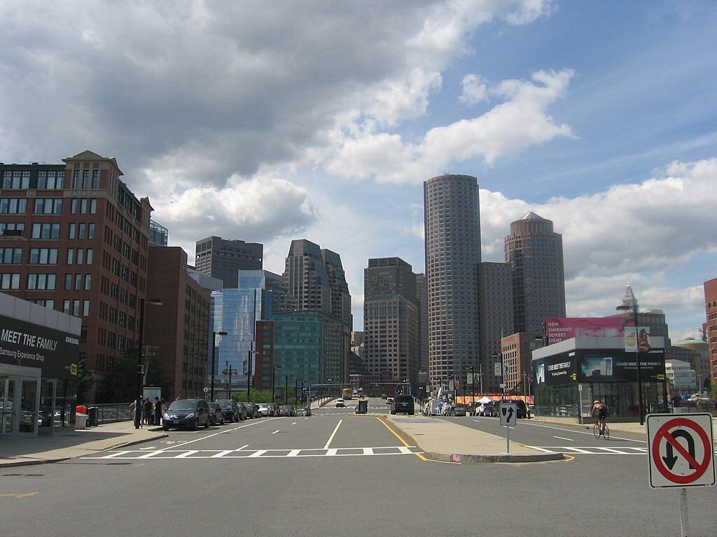 부분적으로 흐린 하늘 아래 높은 건물이 늘어선 도시 거리. 여러 대의 자동차와 보행자가 보입니다.