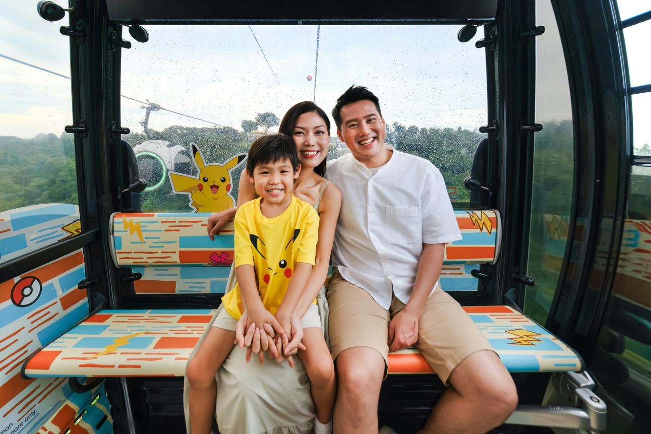 A smiling family of three seated in a decorated cable car featuring Pikachu-themed designs, with greenery visible outside.