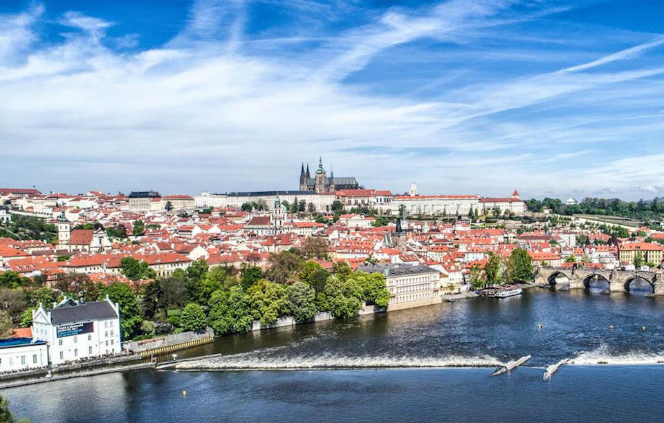 A panoramic view of a city with historic buildings and red roofs by a river, under a blue sky with scattered clouds.