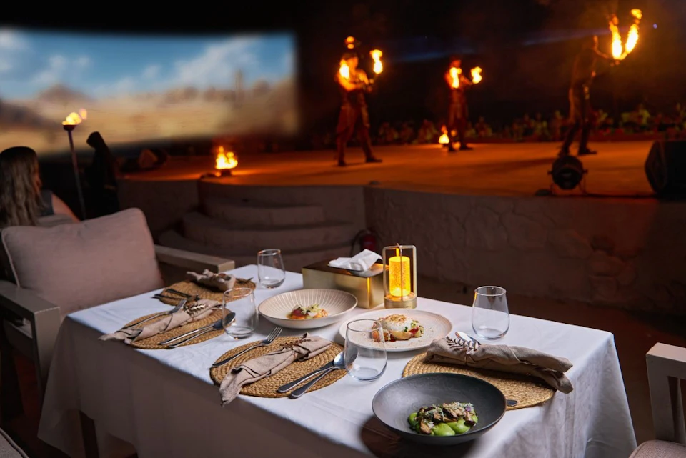 Outdoor dining setup with plates of food on a table in the foreground, and fire performers on stage in the background.