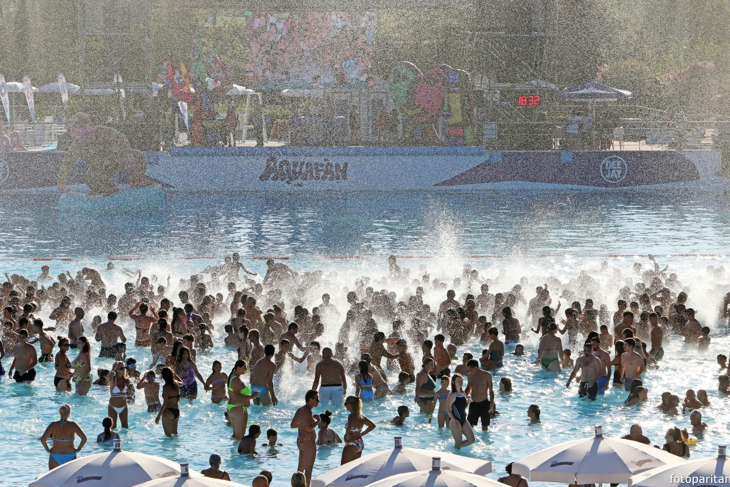 Una piscina all'aperto affollata con molte persone che si godono l'acqua sotto la luce del sole, con lo sfondo di un parco acquatico e gli ombrelloni in primo piano.