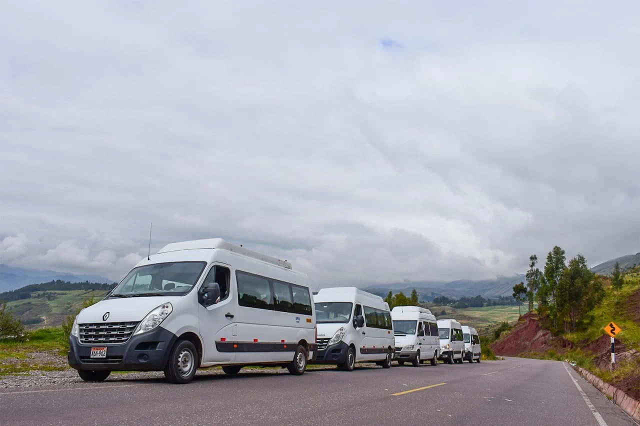 A row of white passenger vans parked on a rural road under a cloudy sky, with grass fields and mountains in the background.
