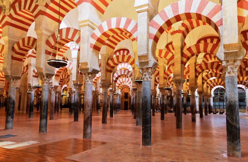 A large hall with numerous columns and red-and-white striped arches, tile flooring, and hanging lamps.