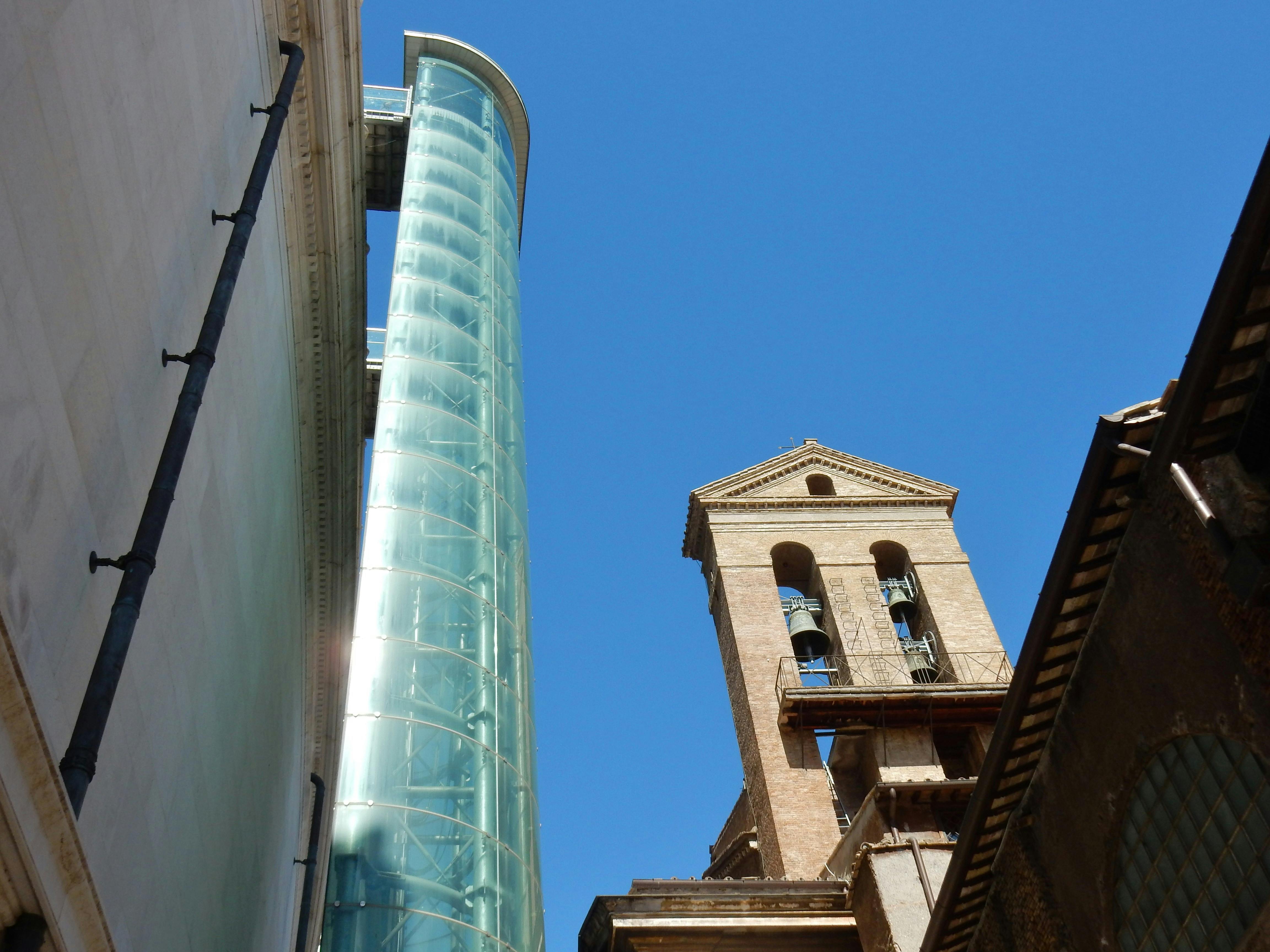澄んだ青い空の下、鐘のある古いレンガの塔の隣には、モダンなガラスのエレベーターシャフトが立っています。