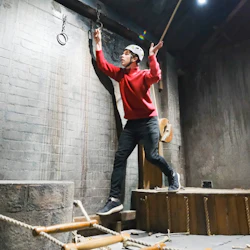 Person i röd tröja och hjälm som sträcker sig efter ringar i ett hinderbanelokal med dödskalle och repstege.