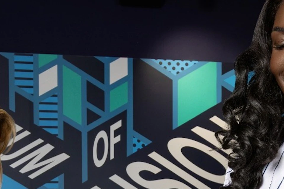 Two people engaging with an interactive exhibit at a museum, smiling and placing objects on a display with a sign in the background.