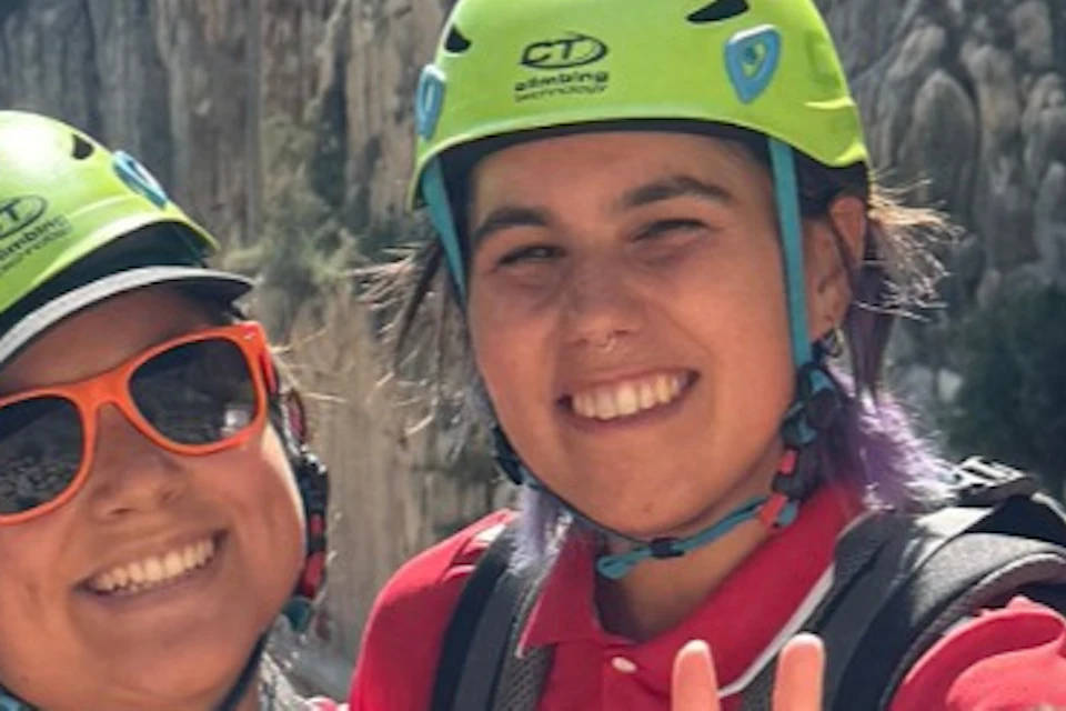 Four people wearing climbing gear and bright green helmets, smiling in front of a rocky cliff under a blue sky.