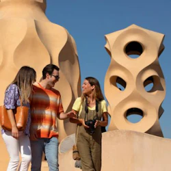 Tre personer, två kvinnor och en man, pratar och ler framför arkitektoniskt unika solbrända byggnader under en klarblå himmel.