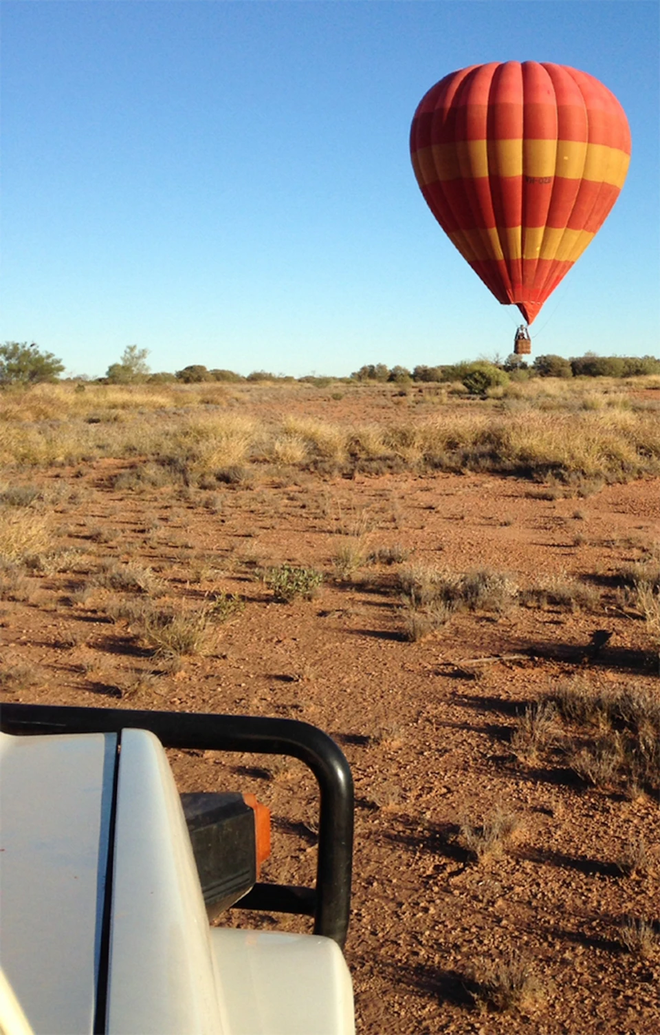 A red and yellow hot air balloon hovers above a dry, grassy plain with sparse vegetation under a clear blue sky.