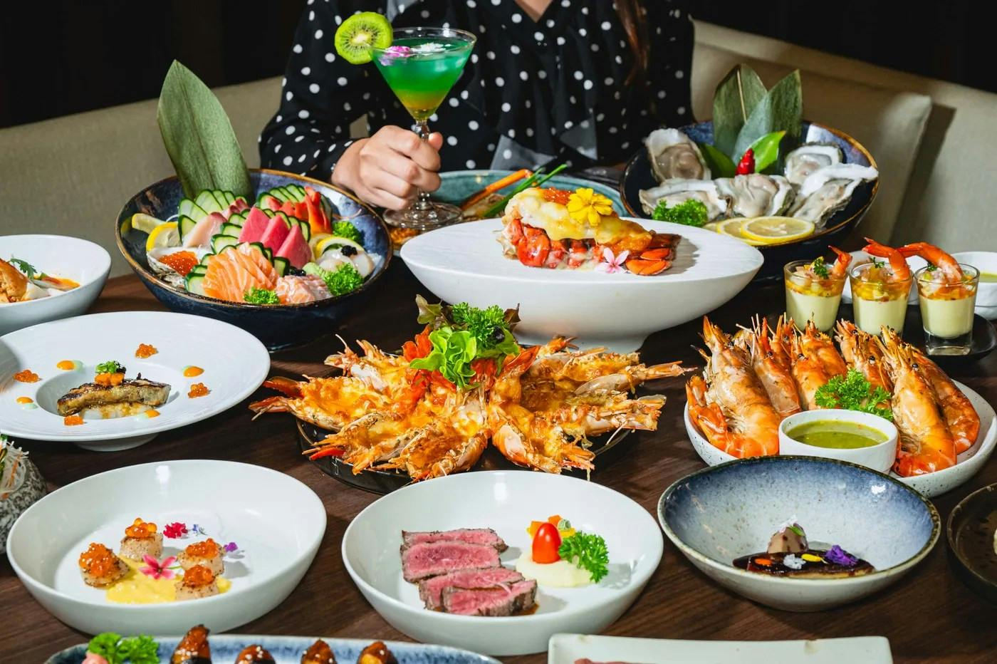A table filled with various seafood and meat dishes, garnished and arranged elegantly, with a person holding a cocktail.