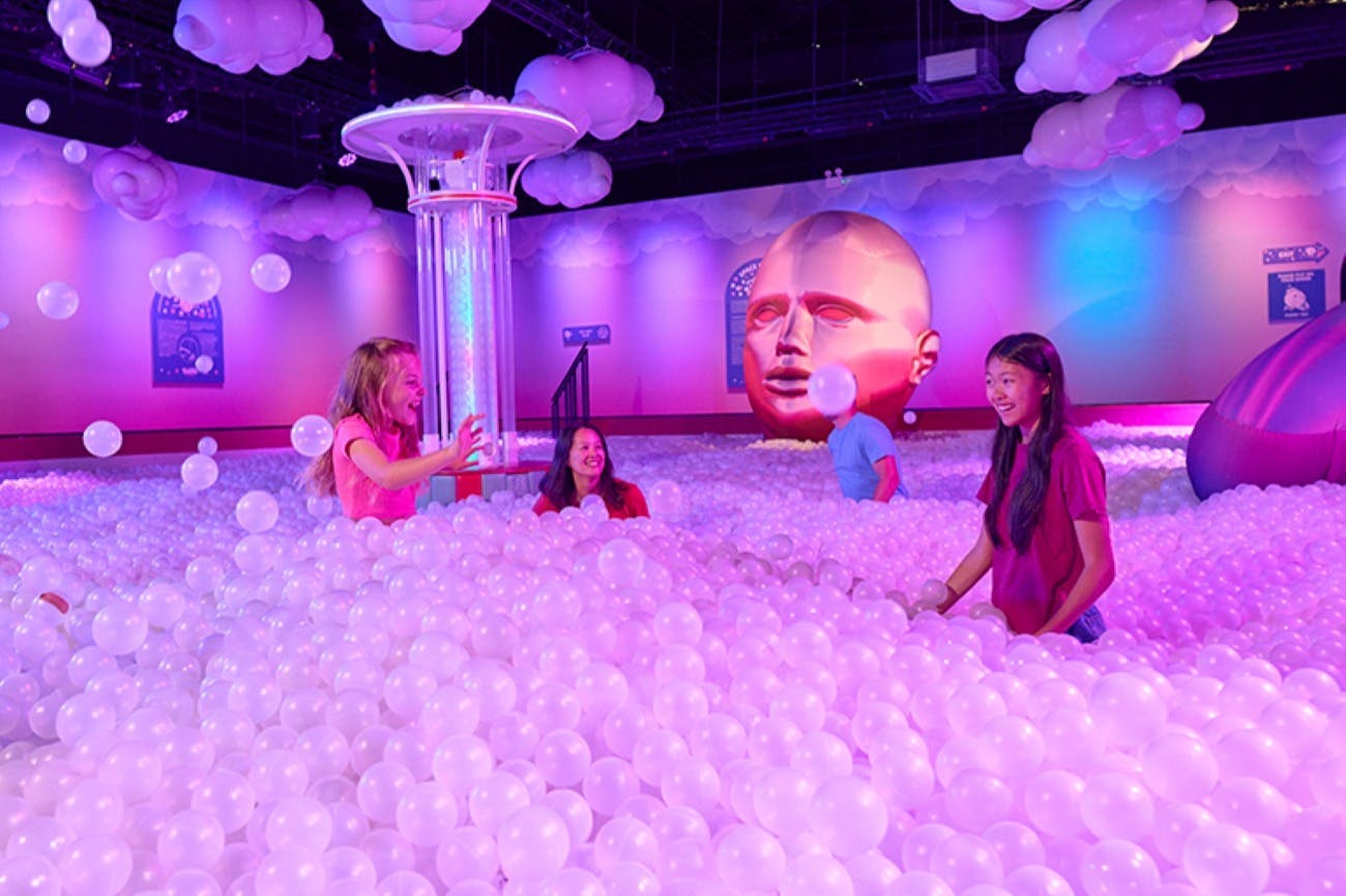 Människor som spelar i en färgglad bollgrop omgiven av nyckfulla dekorationer och en stor upplyst ansiktsskulptur.