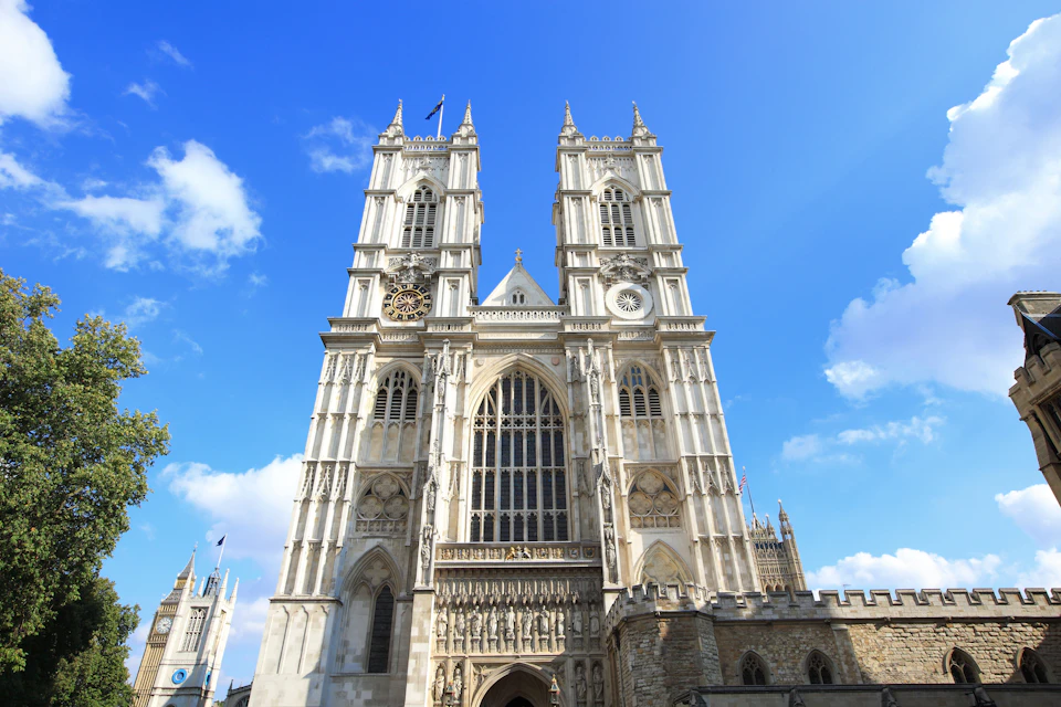 Gothic-style cathedral with twin towers, stained glass windows, clock, detailed stone facade, under a blue sky with scattered clouds.