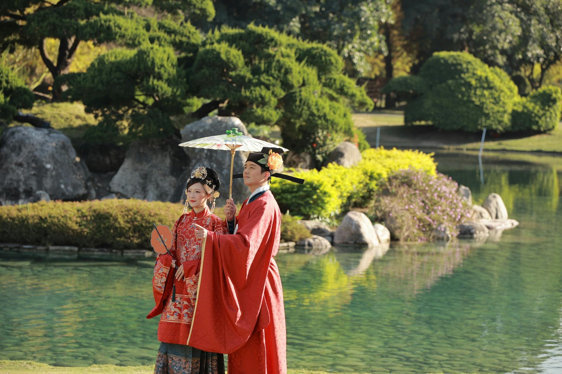 Two people in traditional attire standing near a pond, one holding a decorative umbrella, in a lush garden setting.