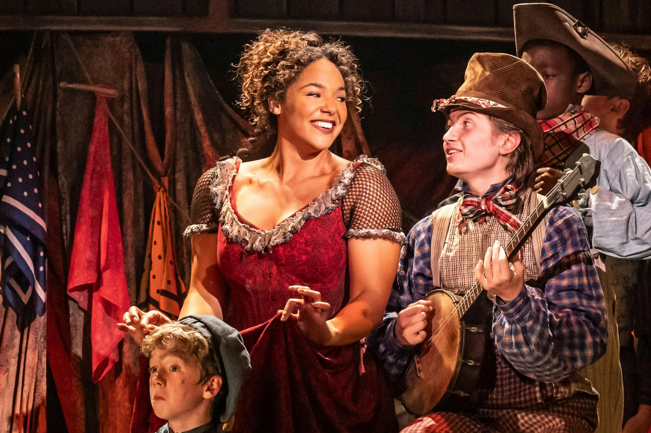 Una dona amb un vestit vermell somriu a un home amb camisa a quadres i barret tocant un banjo. Hi ha altres persones amb disfresses d'època a prop.