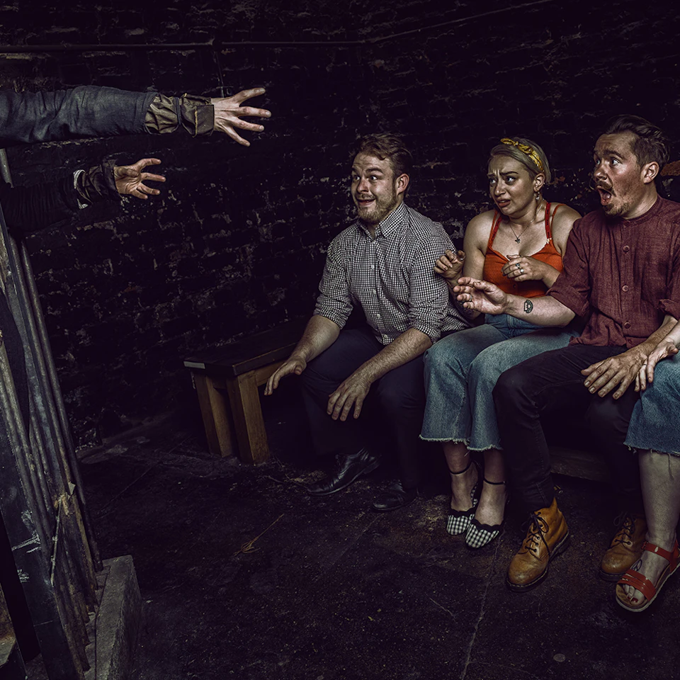 Three people on a bench react with fear and excitement as a person to the left reaches towards them in a dark room.