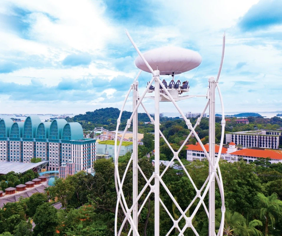 View of a tall observation ride with people seated at the top, surrounded by buildings and lush greenery under a partly cloudy sky.