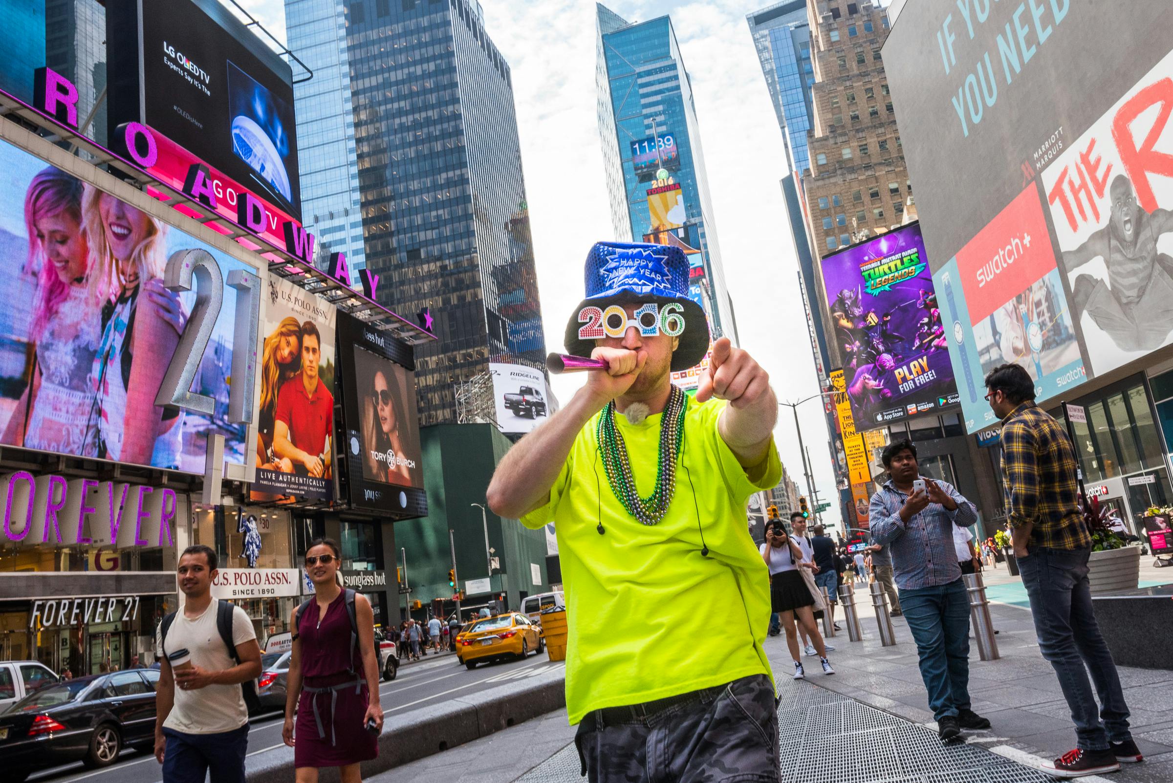 Ένας καλλιτέχνης του δρόμου με φωτεινά ρούχα ποζάρει με ένα μικρόφωνο στην Times Square, περιτριγυρισμένος από διαφημιστικές πινακίδες και πολυάσχολους πεζούς.