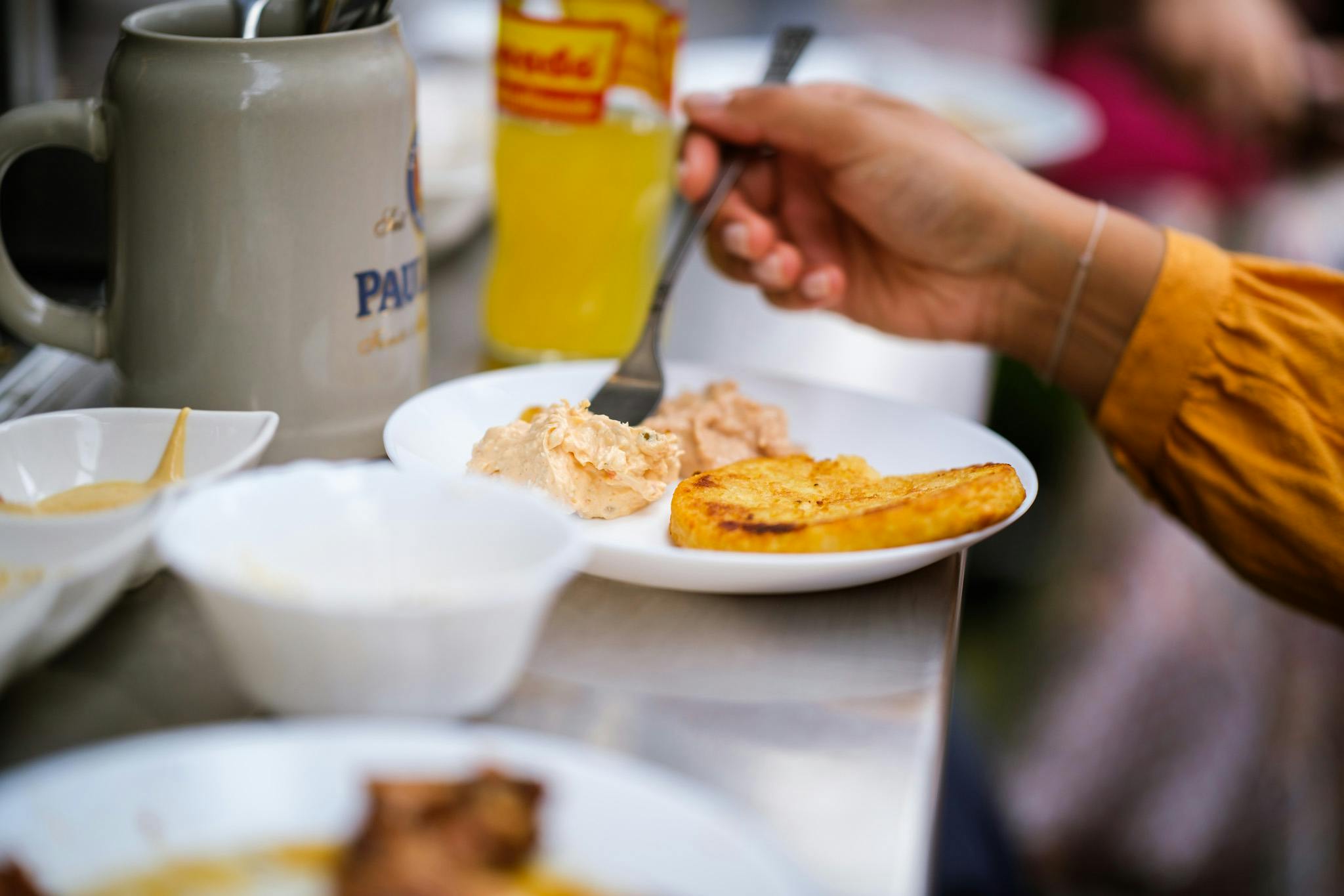 Osoba, která jí jídlo z bílého talíře obsahujícího plátek chleba a pomazánku, s lahví žlutého nápoje v pozadí.