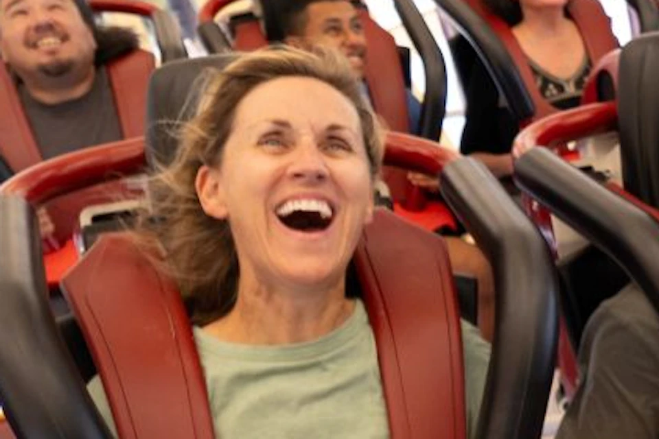 People seated on a roller coaster ride, smiling and enjoying the experience, with a parking lot visible in the background.