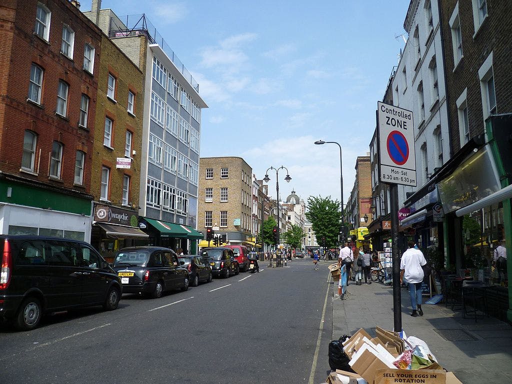 Straße mit beidseitig geparkten Autos, Fußgängern, die an Geschäften vorbeilaufen, einem Schild "Kontrollierte Zone" und Gebäuden entlang der Straße.