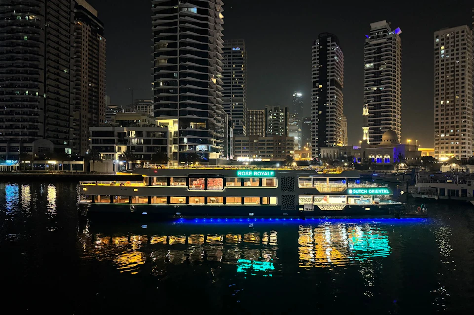 A lit-up cruise boat with signs "Rose Royale" and "Dutch Oriental" on a city waterfront at night, skyscrapers in the background.
