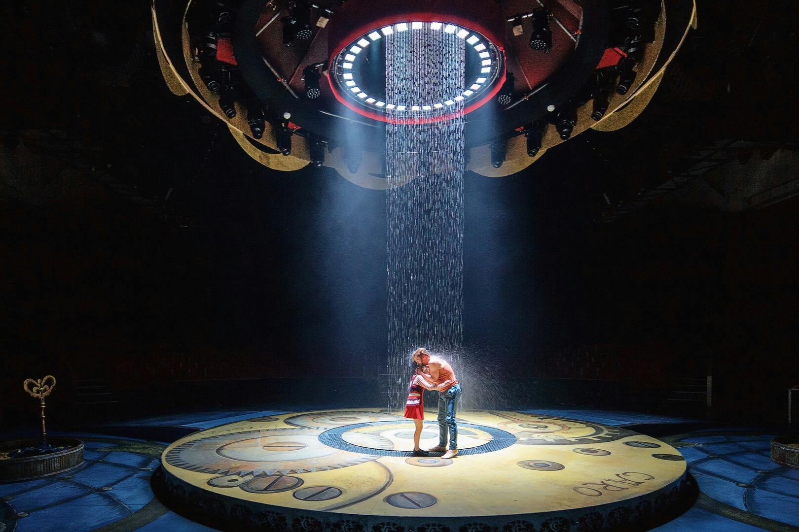 A man and a child stand under a spotlight on a stage with a circular design, surrounded by hanging equipment and dripping water.