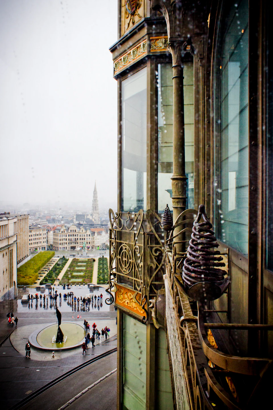 Elevated view of a cityscape with gardens, a spire in the background, and intricate wrought iron details on the right.