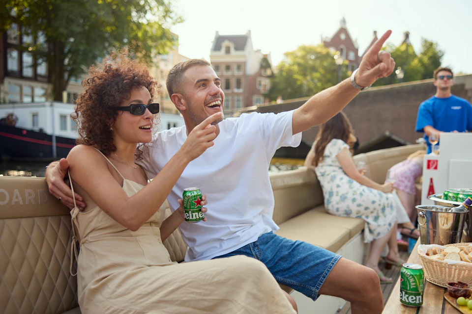 A smiling couple enjoys a boat ride on a sunny day, with canal houses and trees visible in the background.