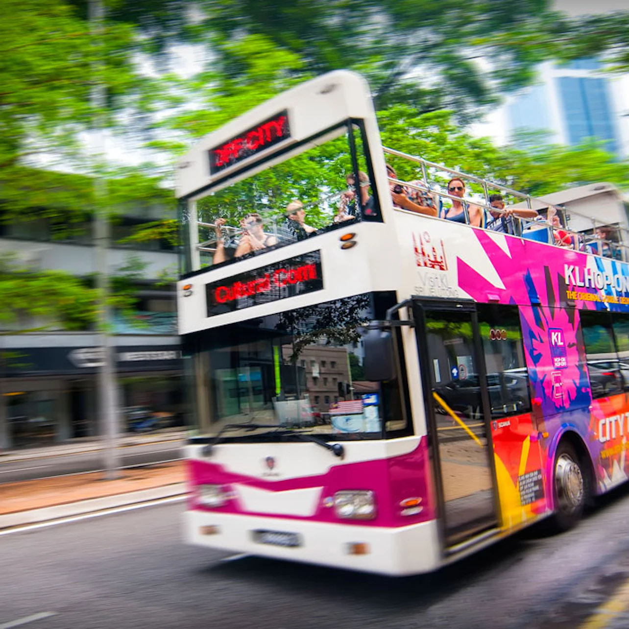 A double-decker bus with vibrant advertisements and tourists on the upper deck travels through a city street lined with trees.