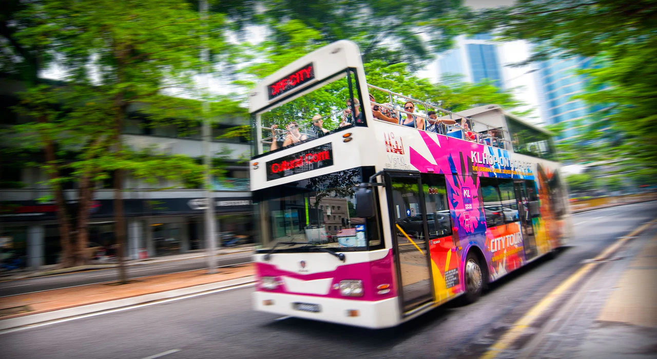 A double-decker bus with vibrant advertisements and tourists on the upper deck travels through a city street lined with trees.