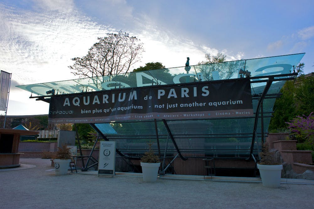 Entrada do Aquarium de Paris com uma faixa preta que indica "não é apenas mais um aquário". Árvores e um céu nublado ao fundo.