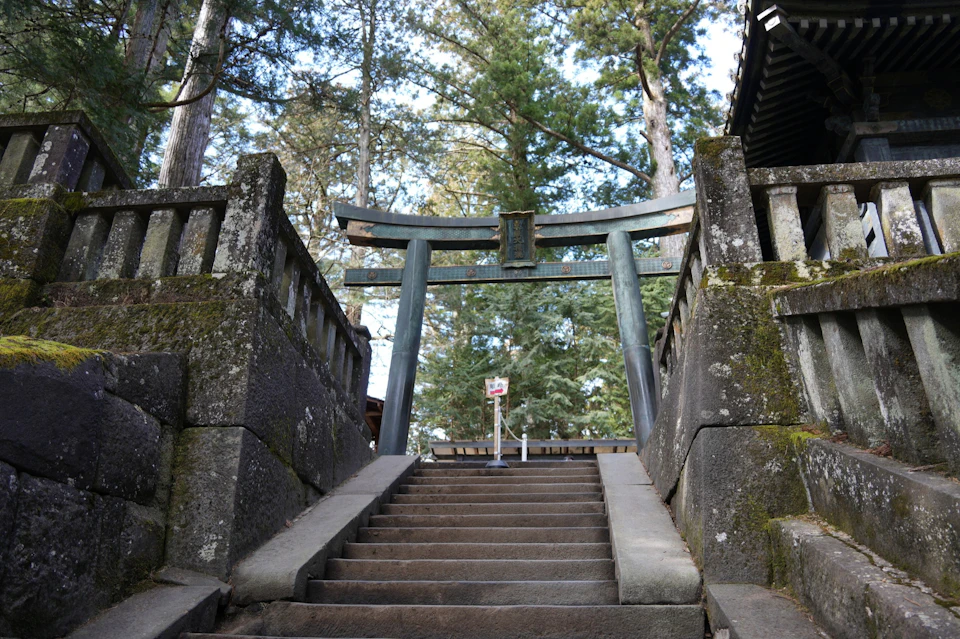 A stone staircase leading to a traditional Japanese torii gate, surrounded by trees and stone walls.