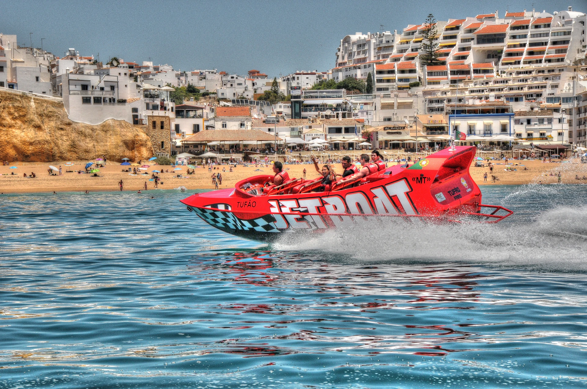 A red jet boat with passengers speeds across the water near a busy beach with buildings in the background.
