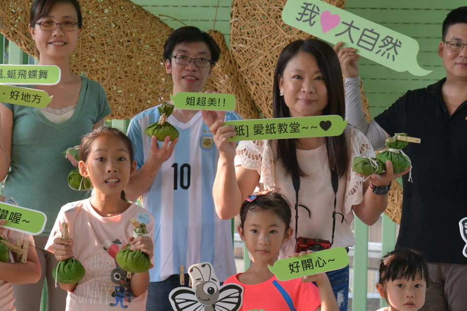 A group of people, including children, holding signs and crafts, standing in front of a green wall with a butterfly decoration.