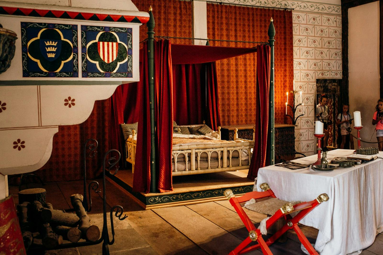 A medieval-style bedroom with a four-poster bed draped in red curtains, a decorated canopy, a fireplace, and a white table with red chairs.
