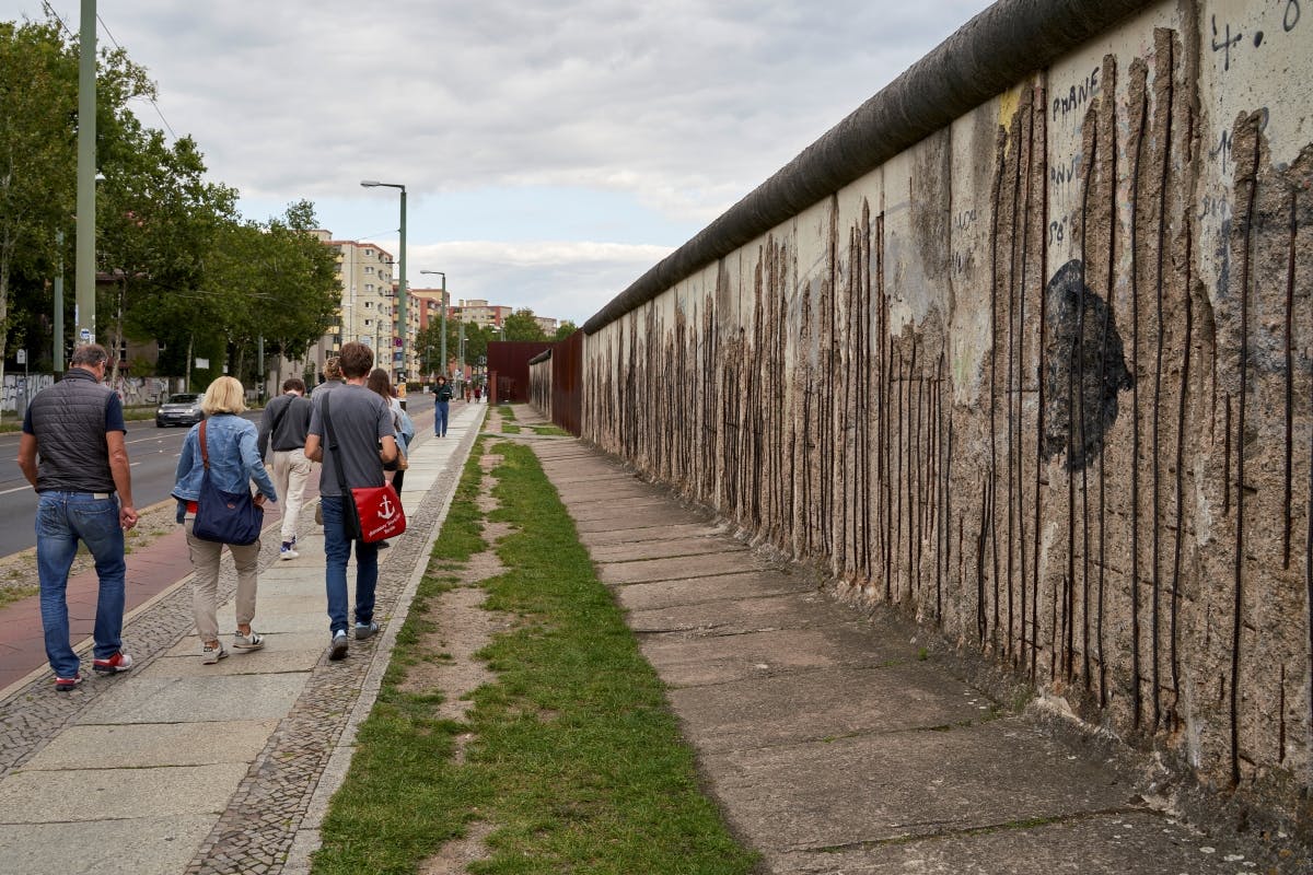Menschen, die unter bewölktem Himmel an einem Stück der Berliner Mauer entlanggehen, mit einer Grasfläche und einem Weg daneben.