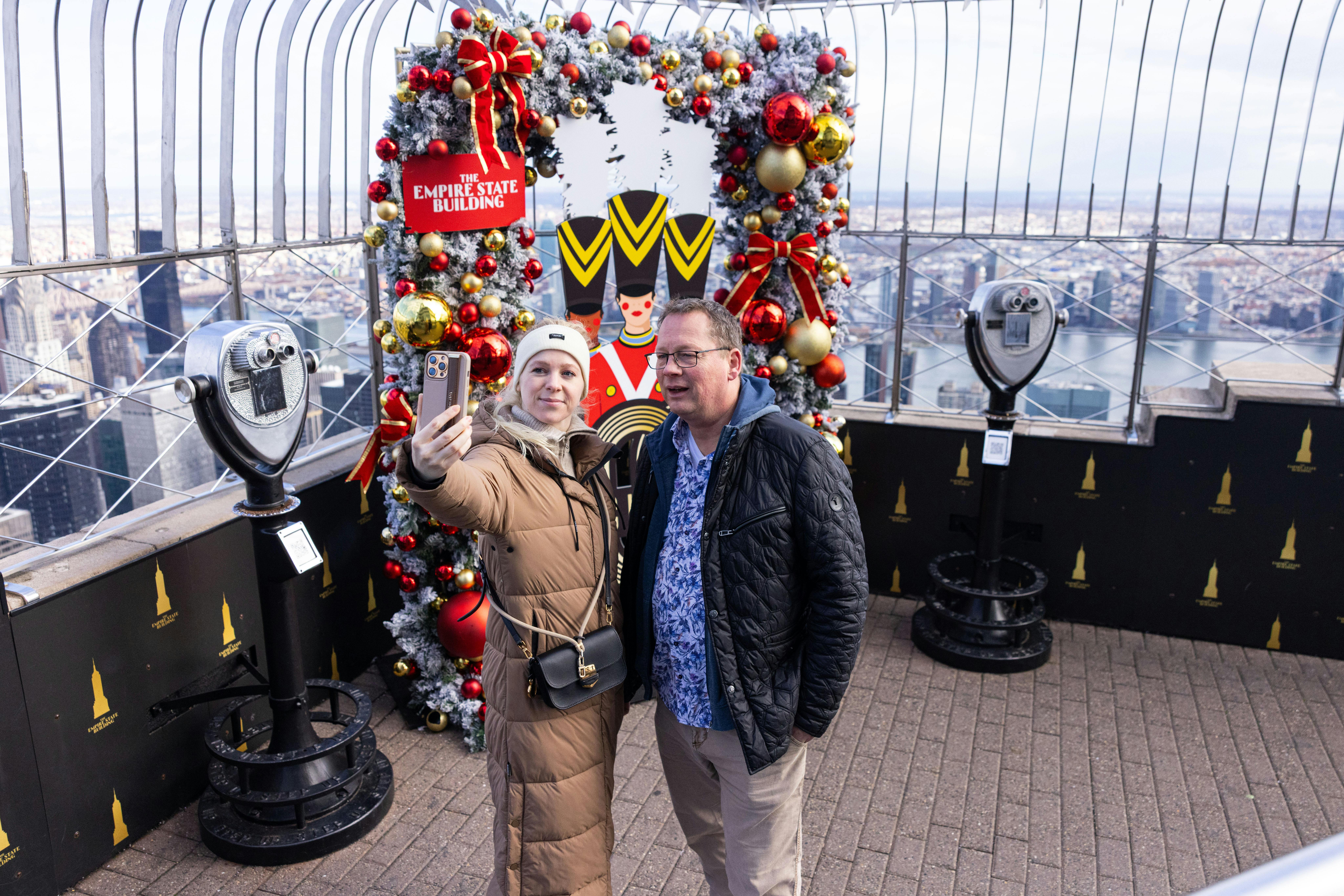 Two people taking a selfie in front of a festive holiday display atop a building, with binocular viewers nearby.