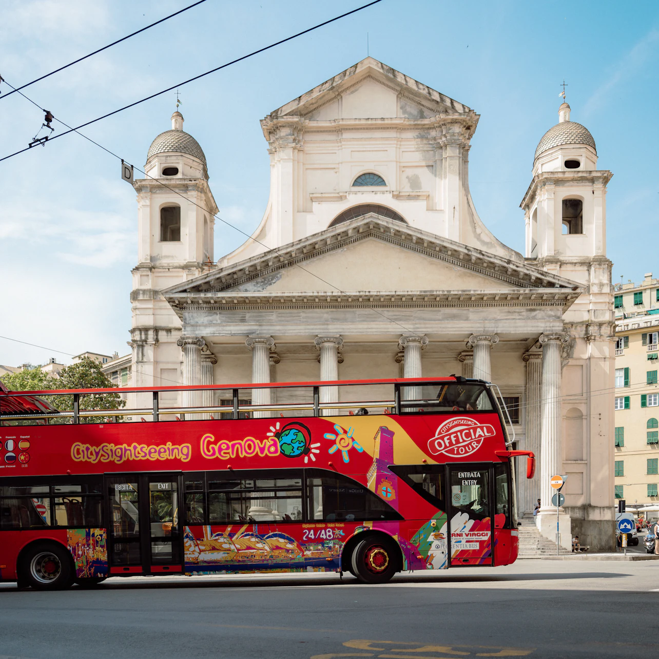 A red sightseeing bus parked in front of a historic church with two bell towers and a blue sky in the background.