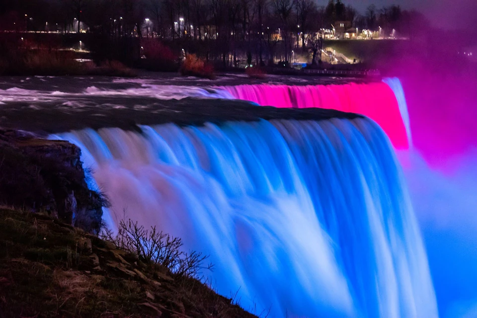 A waterfall illuminated in blue and pink light against a nighttime backdrop with trees and distant city lights.