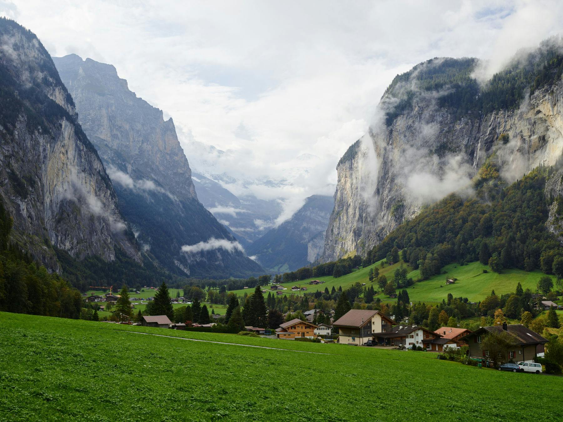 Green valley with scattered houses, surrounded by tall mountains and mist. Cloudy sky overhead.