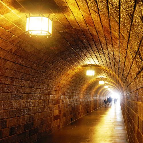 En tegeltunnel upplyst av hängande ljus, med människor som går mot den ljusa utgången längst bort.