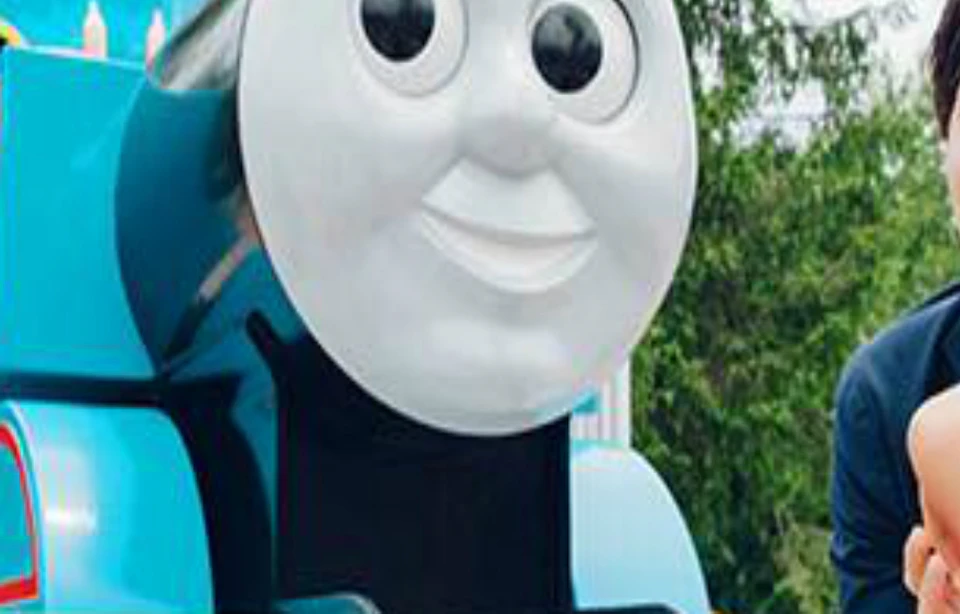 A smiling family poses in front of a large Thomas the Tank Engine model outdoors on a sunny day.
