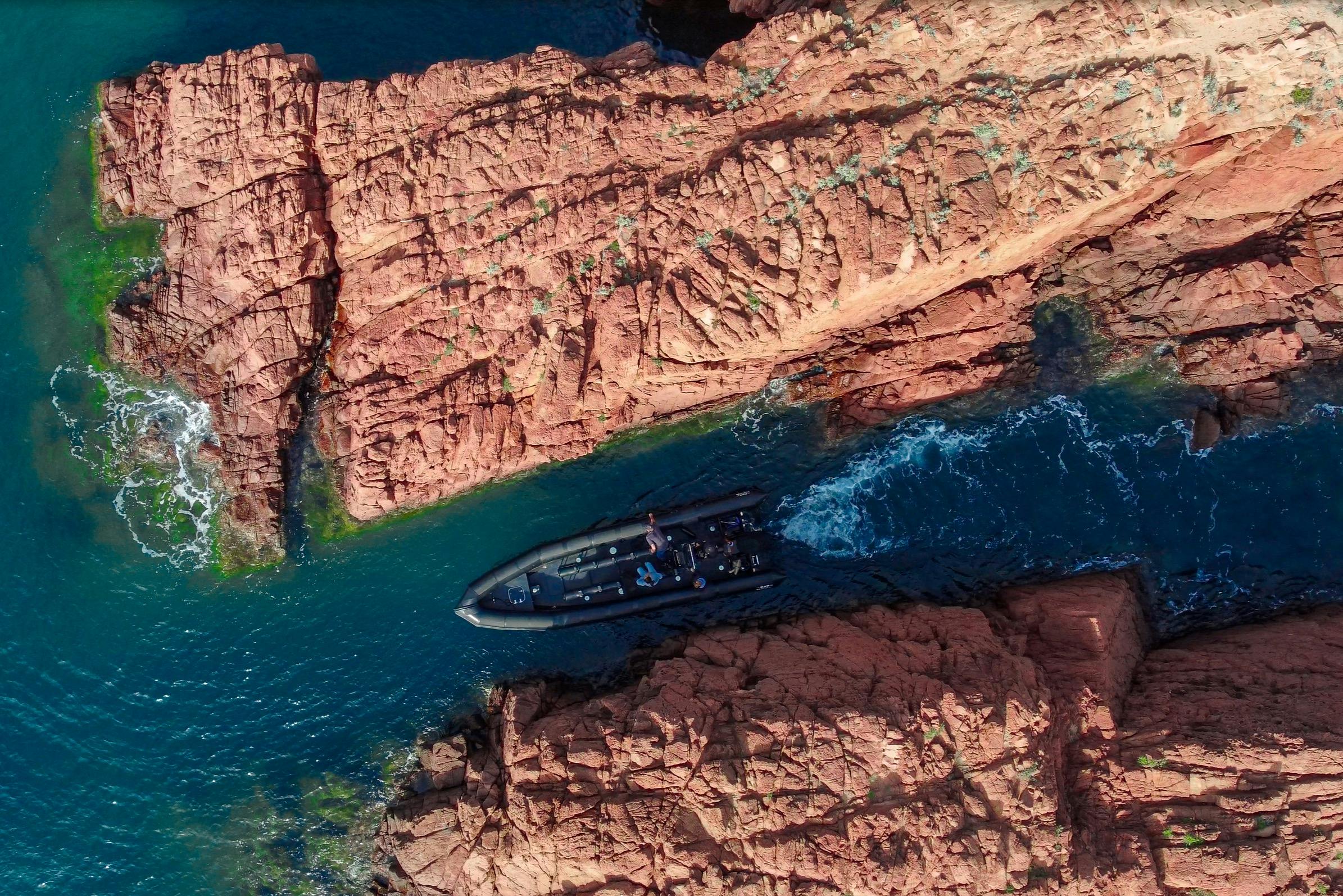 Černá loď proplouvá úzkou vodní cestou obklopenou oranžovými skalními útvary při pohledu shora.
