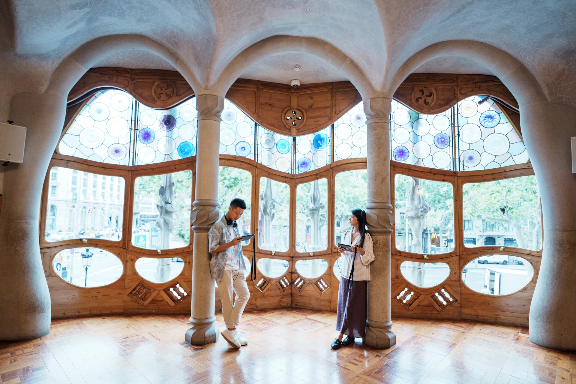 Two people standing in a room with ornate stained-glass windows and wooden decor, holding devices and looking at them.