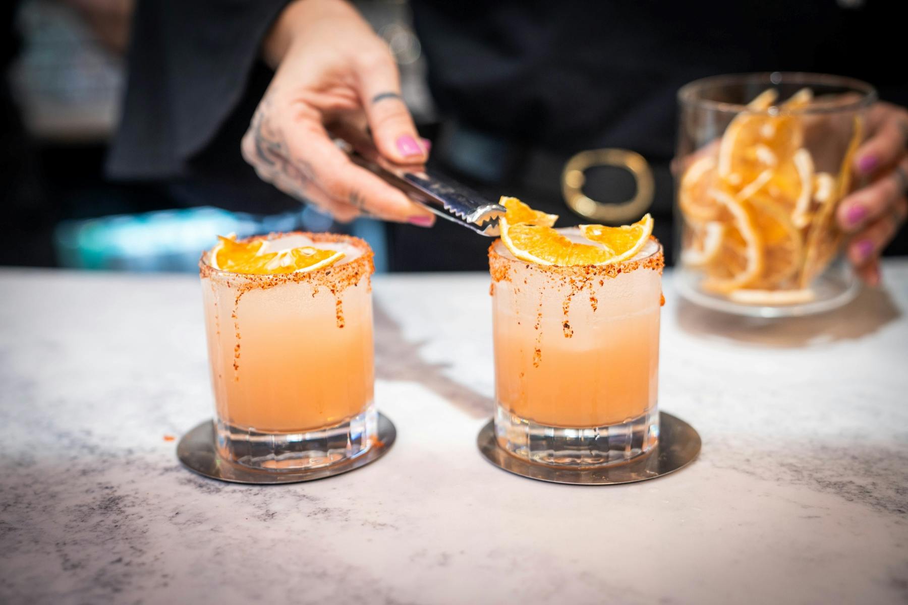 オレンジスライスとシュガーリムの2つのカクテルは、トングを使用して手で飾られています。 オレンジ色のスライスの瓶が背景にぼかされています。