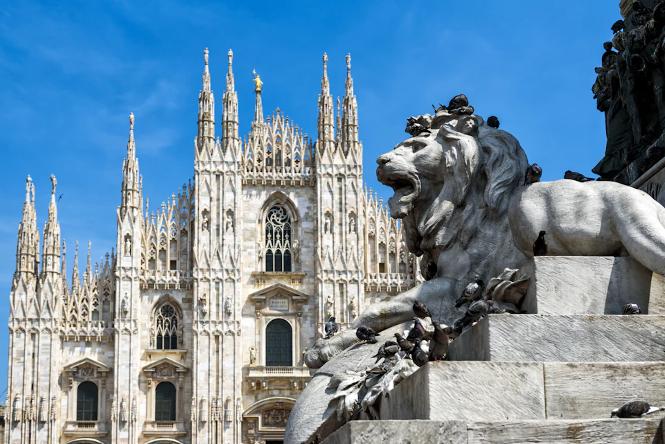 Statue of a lion with pigeons resting on it, set in front of a gothic cathedral with ornate spires against a clear blue sky.
