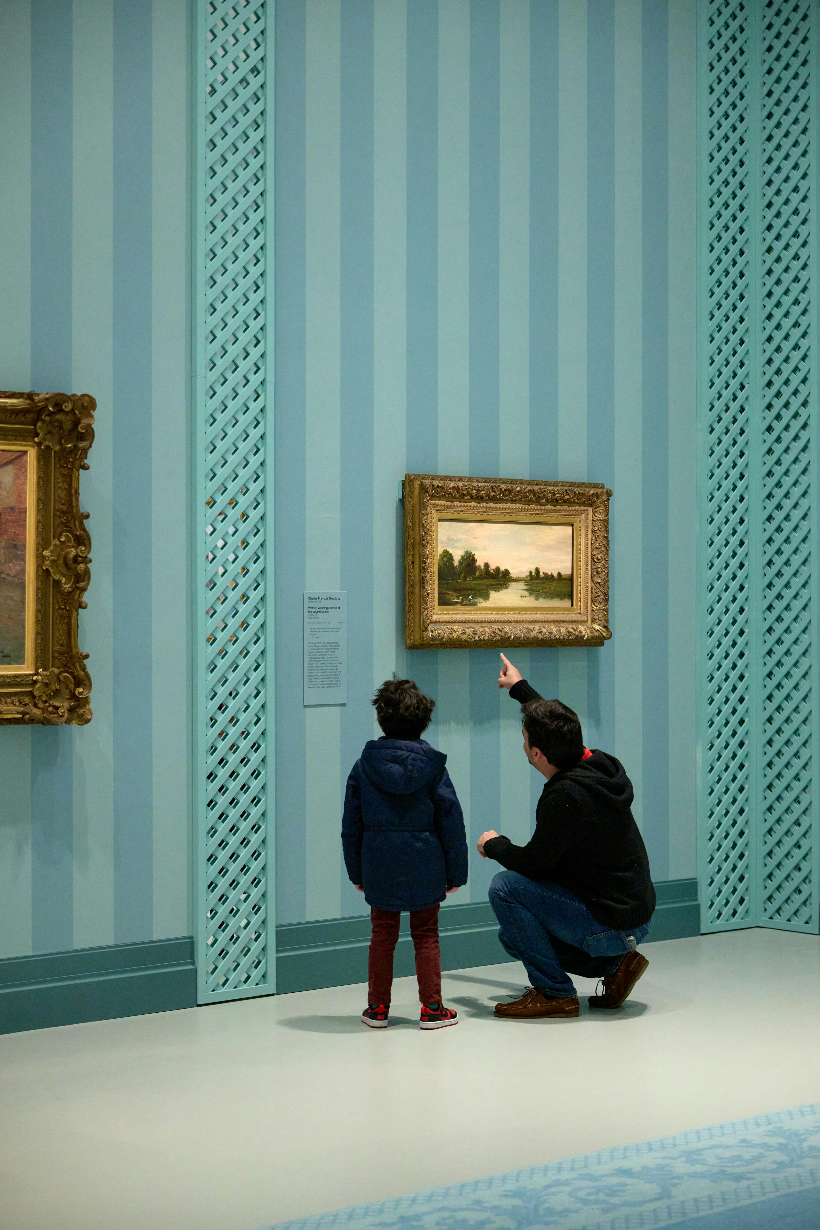 Dospělý klečí a ukazuje na obraz na zdi galerie s modrými pruhy, zatímco dítě stojí vedle nich a pozoruje.