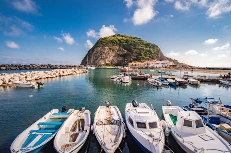 Small boats docked in a marina with a lush, green hill and colorful buildings in the background under a partly cloudy blue sky.