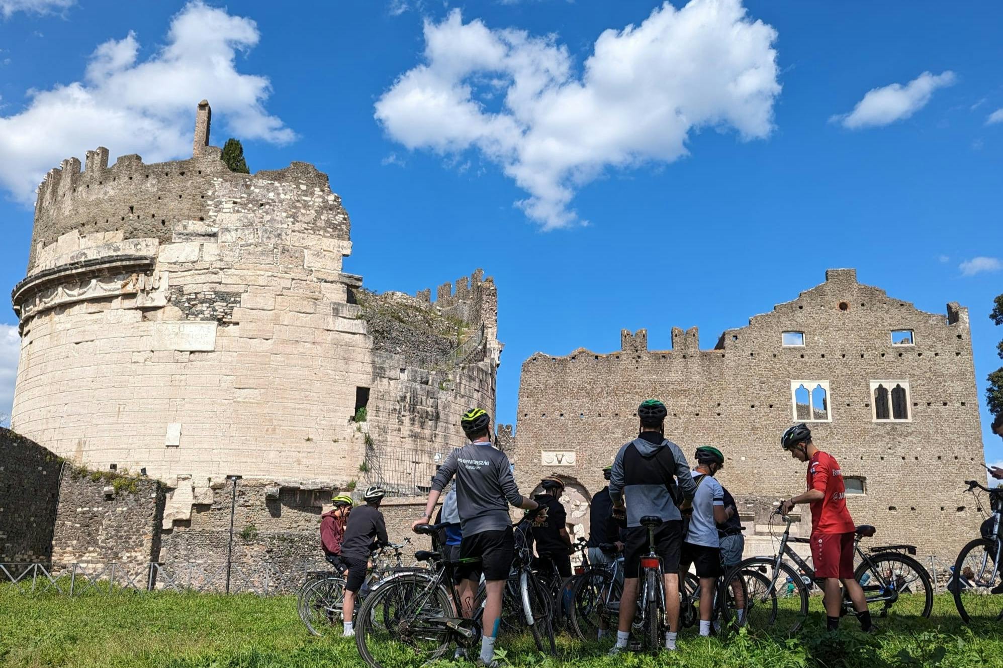 Grupa rowerzystów ze swoimi rowerami zebrała się w pobliżu starożytnych ruin pod błękitnym niebem z rozproszonymi chmurami.