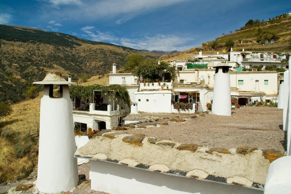 Whitewashed houses with flat roofs and chimneys, set on a hillside with mountains and a blue sky in the background.